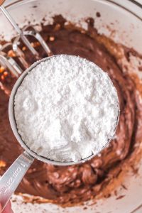 A cup of sifted powdered sugar being added to chocolate butter cream in a bowl - Hostess At Heart
