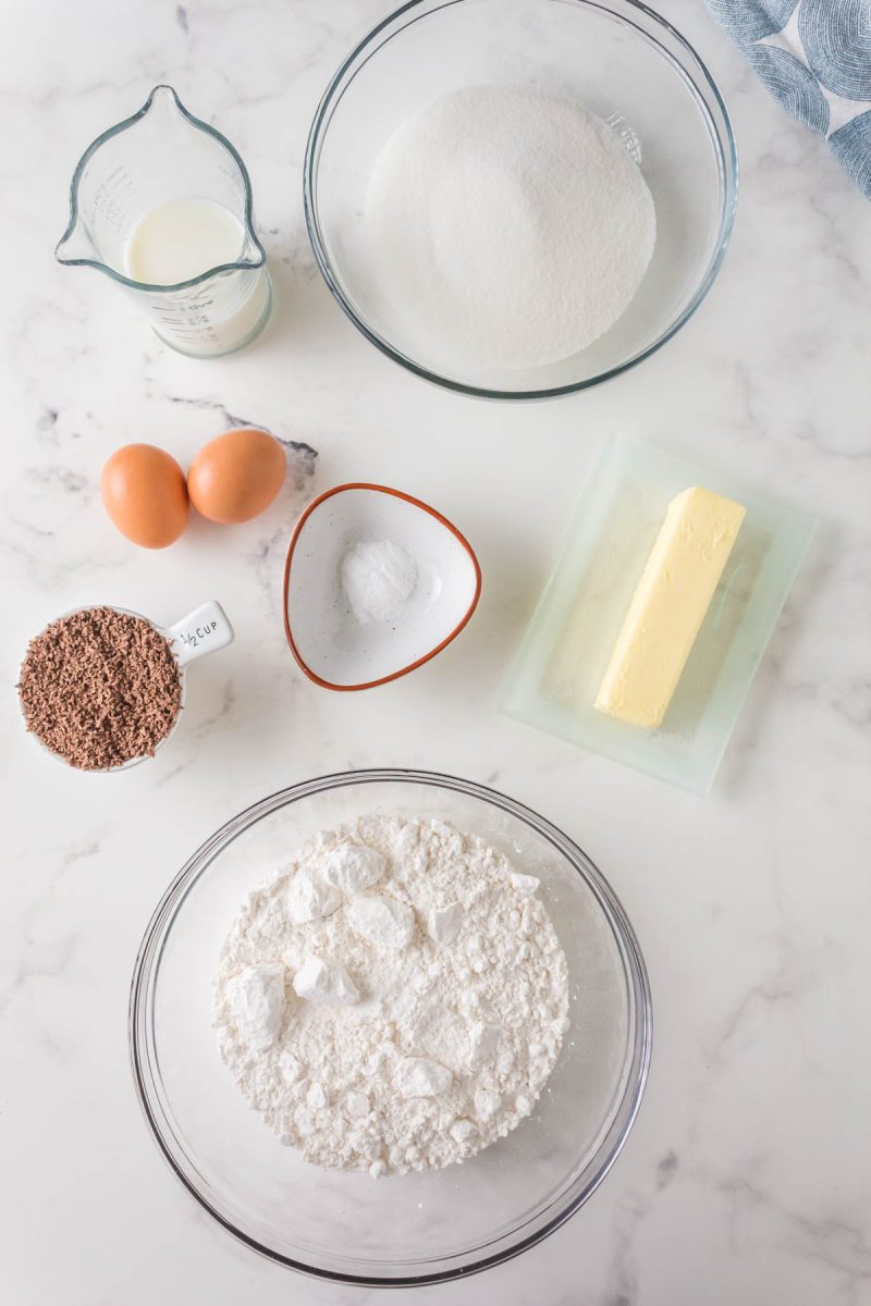 Top down view of ingredients used to make a prize mahogany chocolate cake including flour, butter, sugar, grated chocolate, eggs, milk, and baking soda