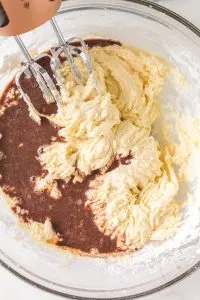 Melted chocolate is added to creamed butter mixture in a bowl.