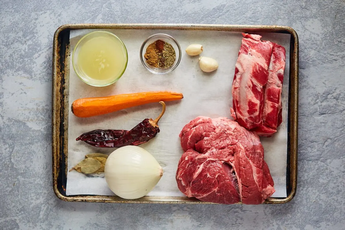 Top down view of beef, onion, garlic, dried pepper, spices, and garlic sitting on a baking sheet. Hostess At Heart