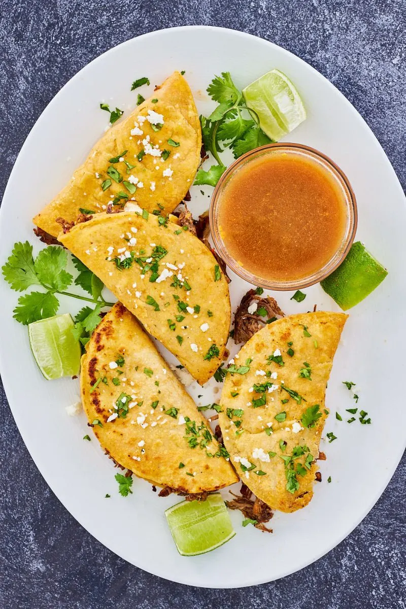 Top-down view of 4 birria meat tacos on a plate garnished with lime wedges, cilantro, and cheese - Hostess At Heart