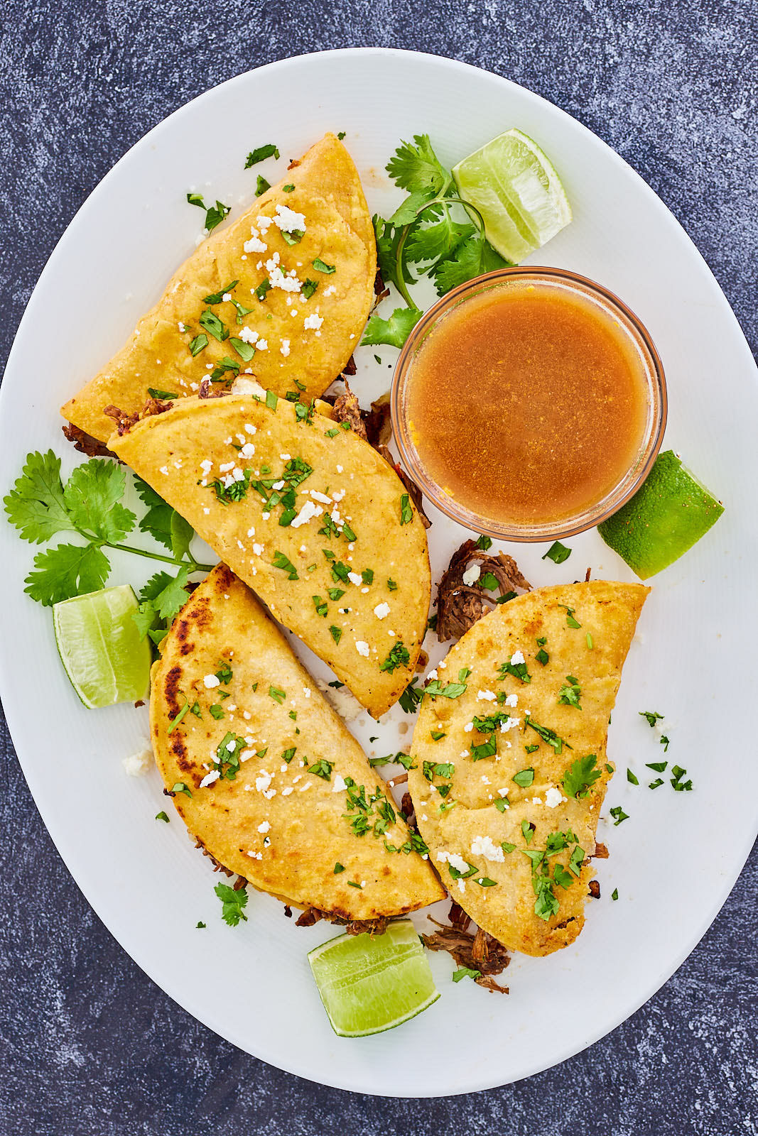 Top-down view of 4 birria meat tacos on a plate garnished with lime wedges, cilantro, and cheese - Hostess At Heart