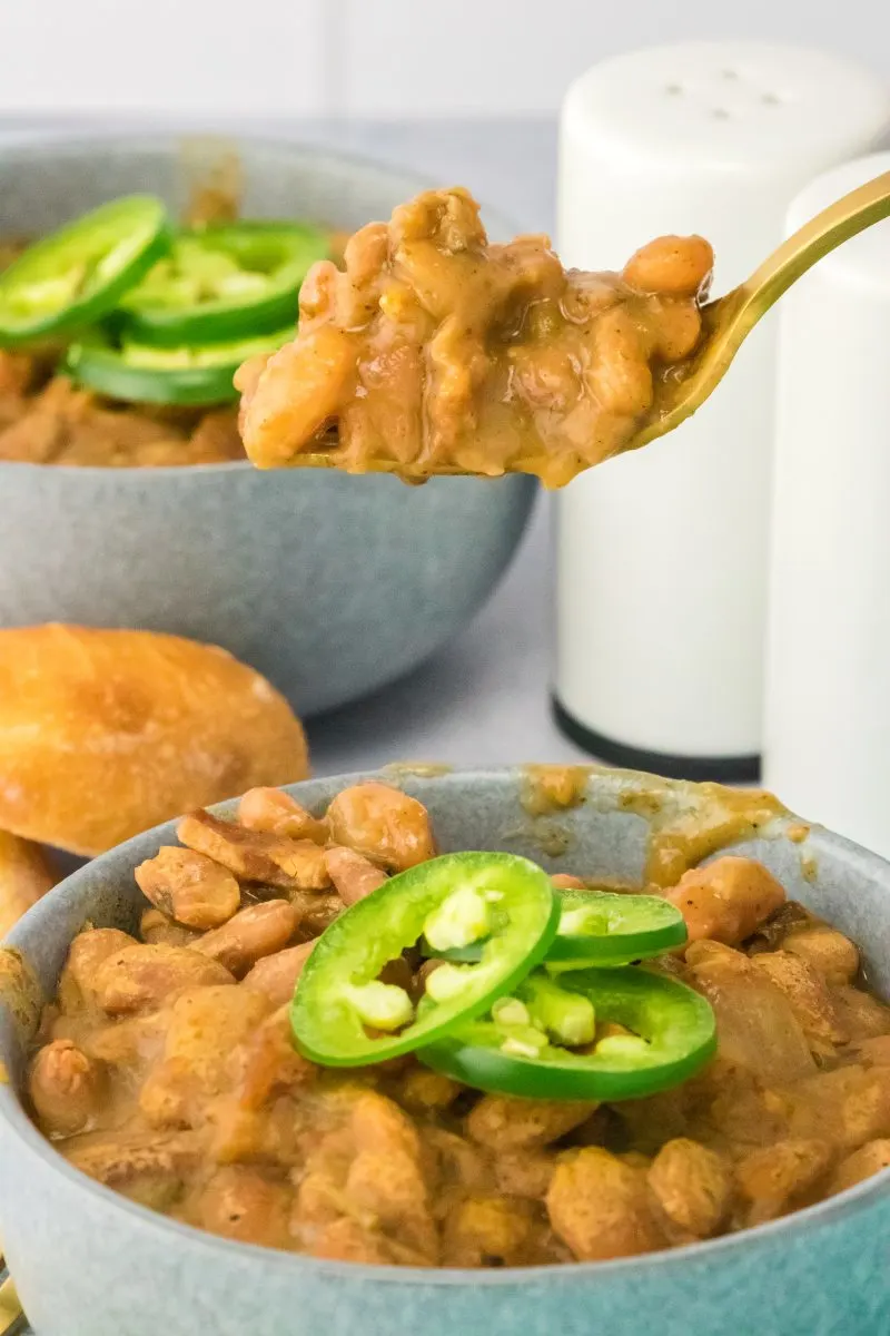 A spoon filled with Borracho beans hovering over a bowl filled with the beans topped with jalapeno slices - Hostess At Heart