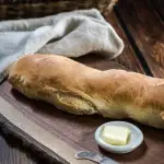 Sideview of a loaf of Italian baguette sitting on a cutting board with a pad of butter next to it.