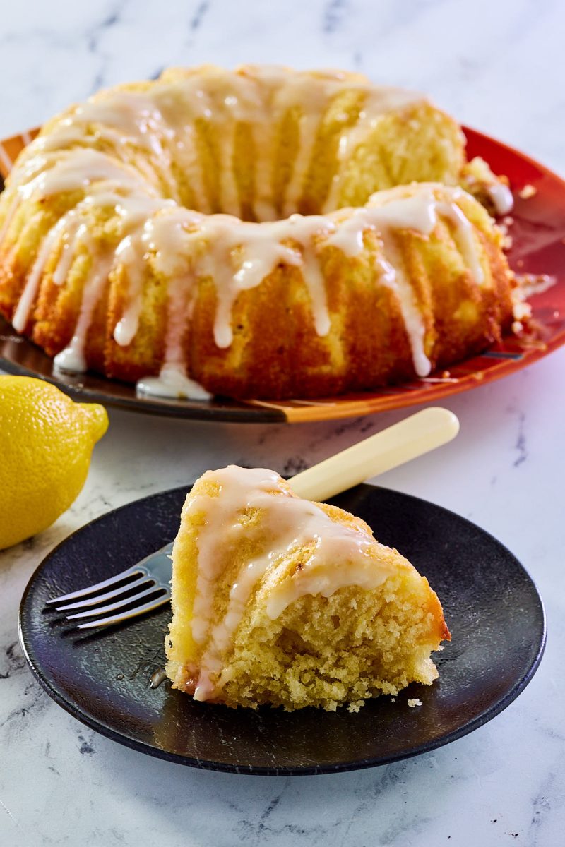 Side view of a slice of Lemon Olive Oil Cake recipe in front of a sliced bundt cake. Hostess At Heart