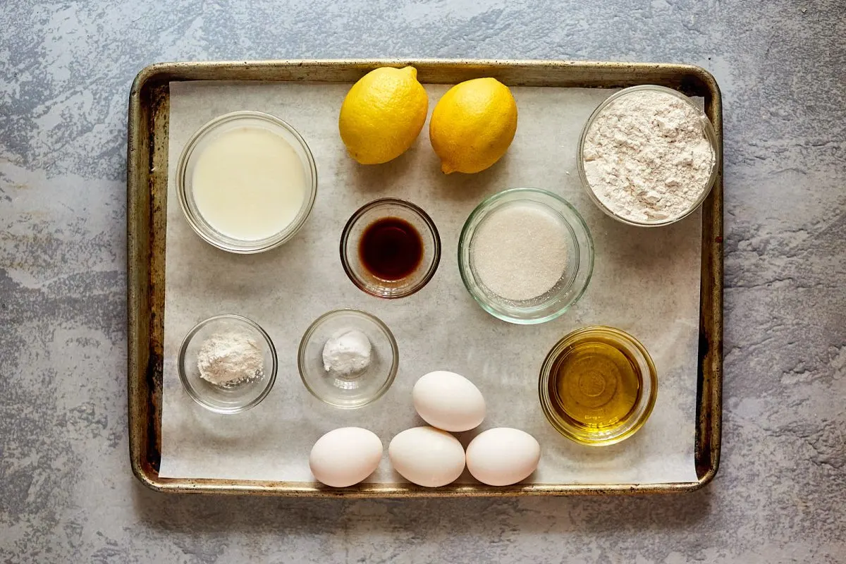 Baking sheet holding dishes of the ingredients used to make a lemon olive oil cake recipe including lemons, flour, sugar, eggs, vanilla extract, baking powder and soda, olive oil, and milk. Hostess At Heart