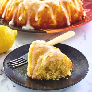 Side view of a slice of glazed lemon olive oil cake sitting on a plate with a bundt cake sitting behind it.