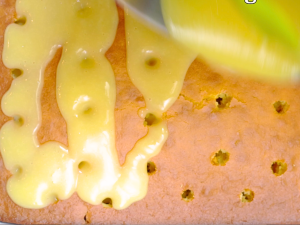 Top-down view of a baked lemon cake poked with holes and a lemon glaze being poured over the top - Hostess At Heart