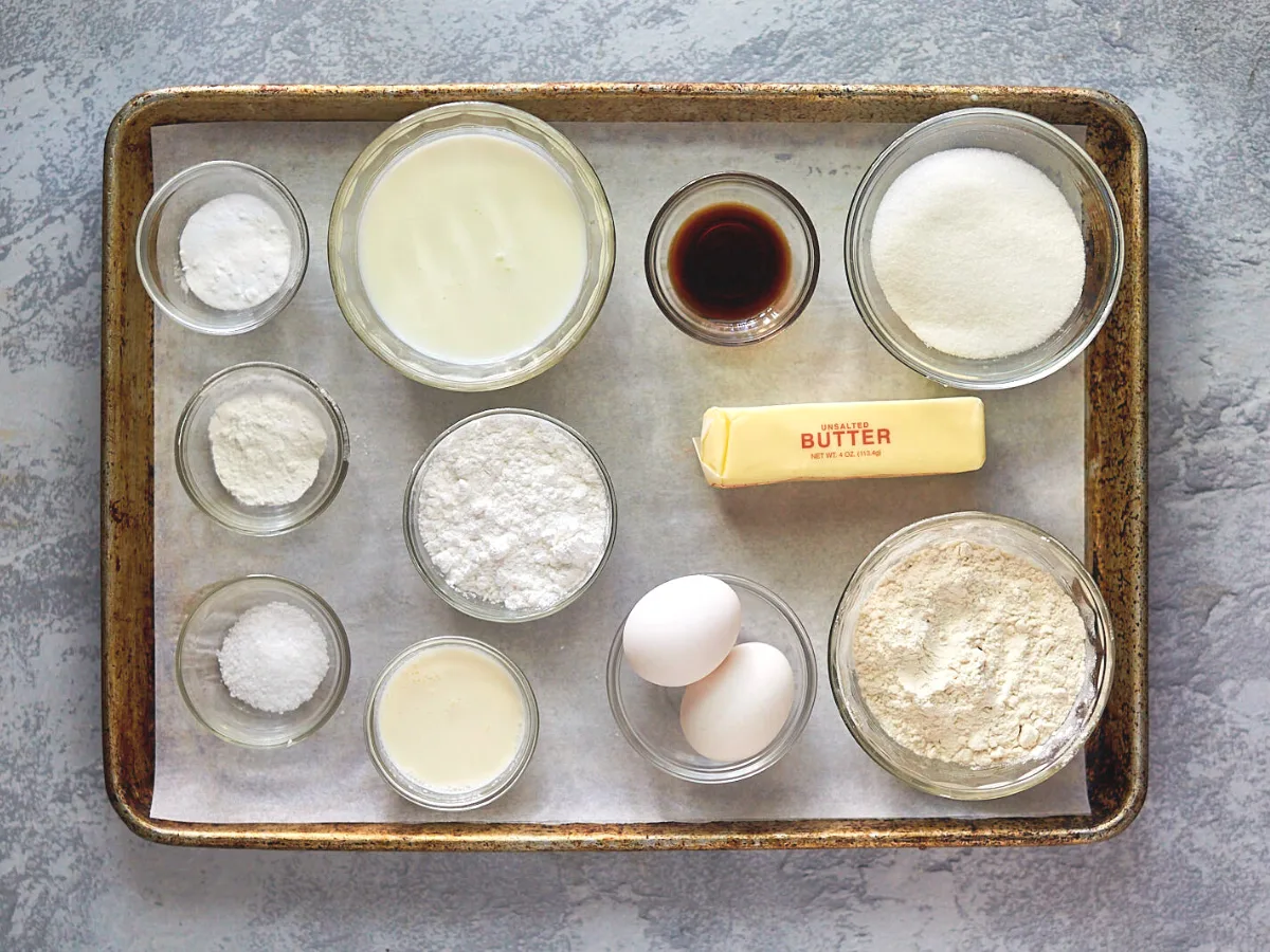 Ingredients for a vanila cake sitting on a baking sheet including buttermilk, sugar, cake flour, eggs, salt, vanilla, baking powder, and baking soda.