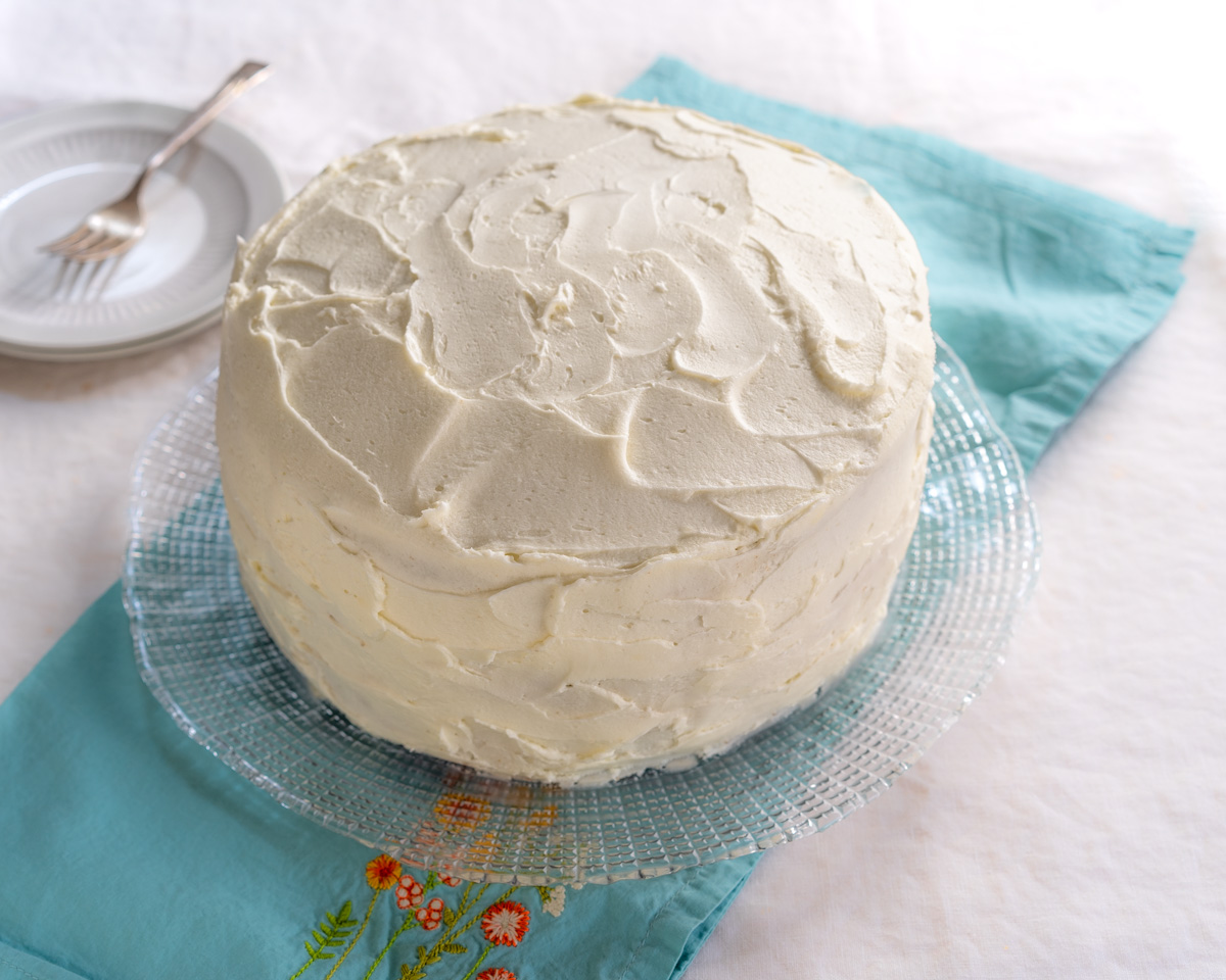 Angled view of a frosted vanilla layer cake sitting on a serving platter - Hostess At Heart