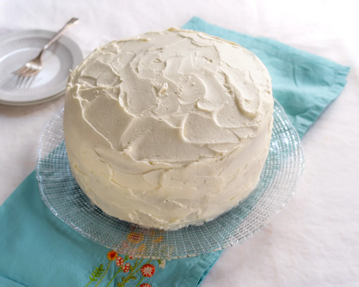 Angled view of a frosted vanilla layer cake sitting on a serving platter - Hostess At Heart