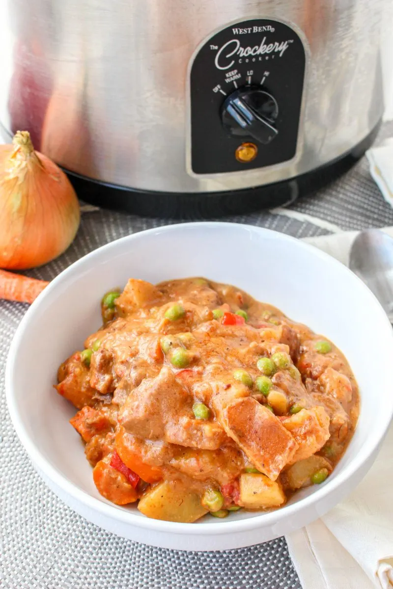 Angled view of a bowl of Caldareta Beef Stew in a bowl with a slow cooker in the back ground - Hostess At Heart