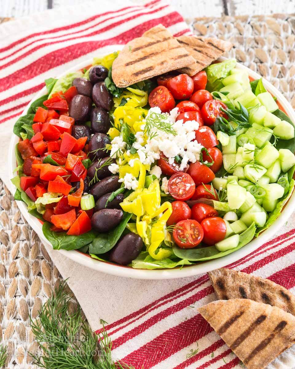 Angled view of a beautiful Greek Salad topped with chopped tomatoes, kalamata olives, peppers, cucumbers, and feta cheese. Grilled pita squares garnish the dish. Hostess At Heart