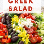 Close-up view of a Mediterranean salad topped with olives, tomatoes, cucumbers, tomatoes, and banana peppers. Hostess At Heart