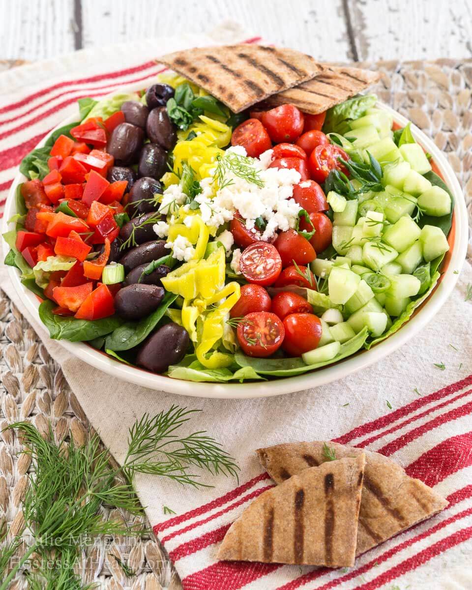 Angled view of a colorful Greek salad topped with chopped tomatoes, cucumbers, olives, peppers, and feta cheese, garnished with grilled pita chips. Hostess At Heart