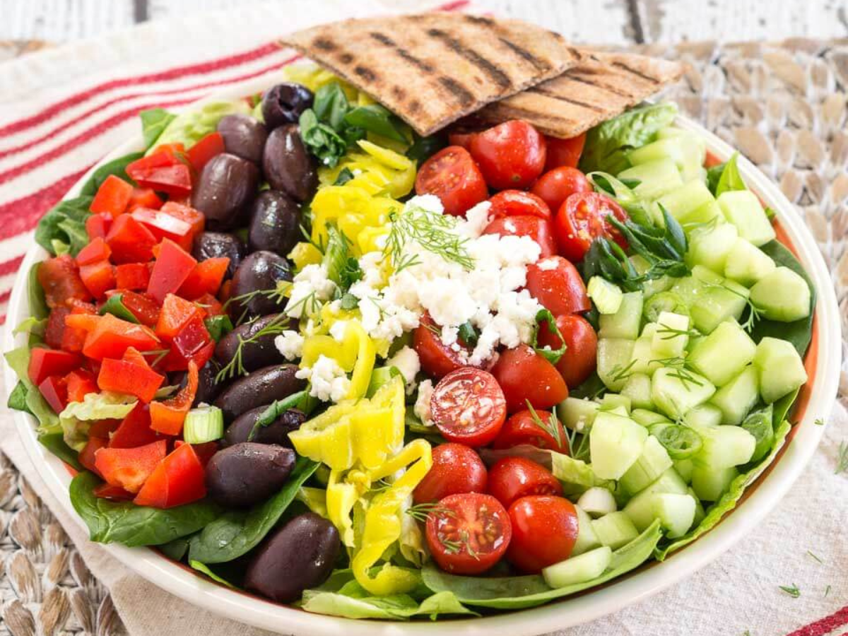Angled view of a large lettuce salad topped with chopped Greek ingredients including peppers, kalamata olives, tomatoes, cucumbers, and Feta Cheese - Hostess At Heart