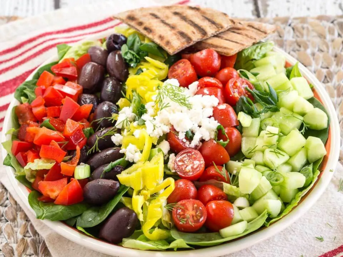 Angled view of a large lettuce salad topped with chopped Greek ingredients including peppers, kalamata olives, tomatoes, cucumbers, and Feta Cheese - Hostess At Heart
