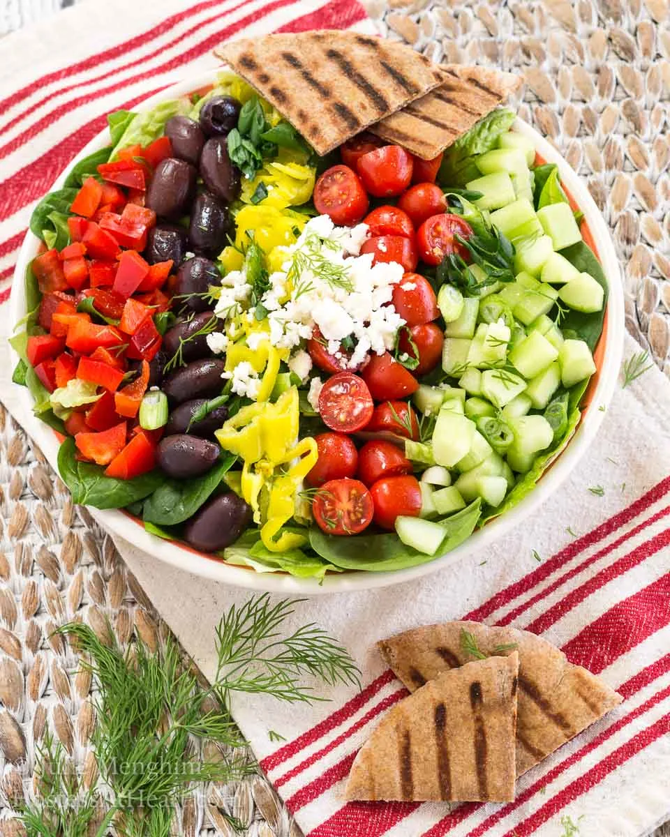 Top-down view of a bowl of lettuce topped with fresh tomatoes, cucumbers, olives, peppers, and Feta cheese and a garnish of fresh dill- Hostess At Heart