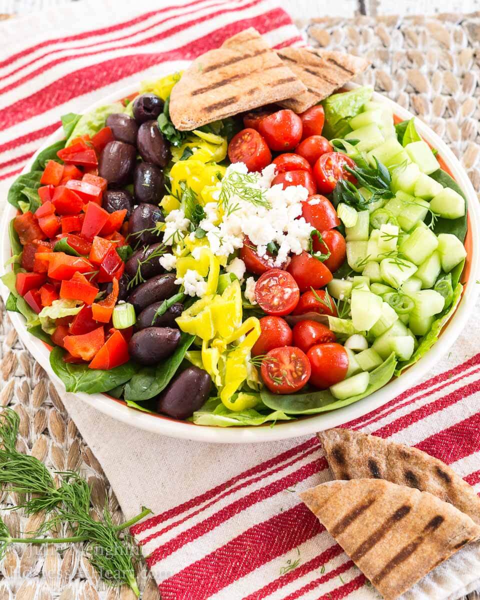 Top down view of a fresh salad topped with healthy ingredients like cucumbers, kalamata olives, onion, Feta, tomatoes, and Greek salad dressing.
