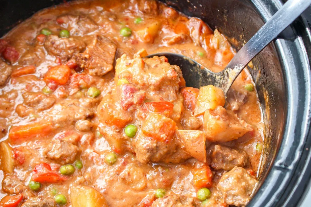Beef and vegetable Caldareta stew in a slow cooker with a serving spoon lifting some out for a closeup view - Hostess At Heart
