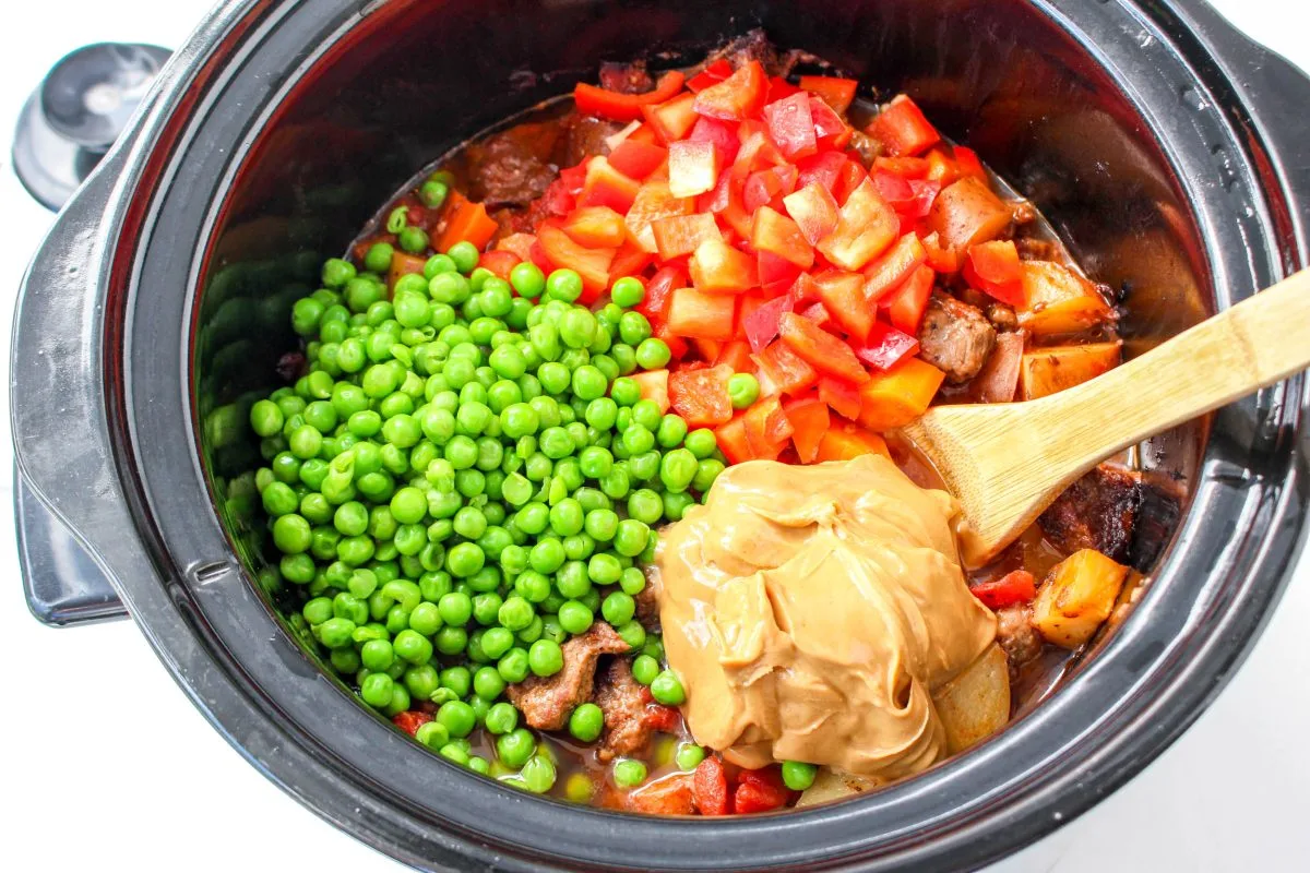 Top down view of a slow cooker with a layer of fresh peas, red peppers, and peanut butter on top of kaldareta stew.