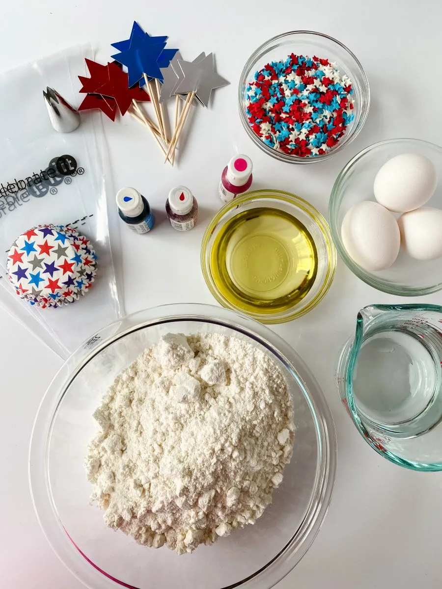 Top-down view of the ingredients needed to make 4th of July cupcakes including cake mix, eggs, oil, food coloring and 4th of July Cupcake decorations - Hostess At Heart