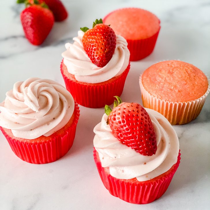 Angled view of freshly baked strawberry cupcakes. Some frosted and topped with a strawberries. Others left plain or just frosted - Hostess At Heart