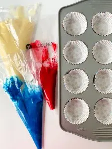 The pastry bags filled with blue, red, and white cupcake batter next to a cupcake pan with liners - Hostess At Heart