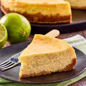 Sideview of a slice of keylime cheese cake with two limes and the whole cheesecake in the back. - Hostess At Heart