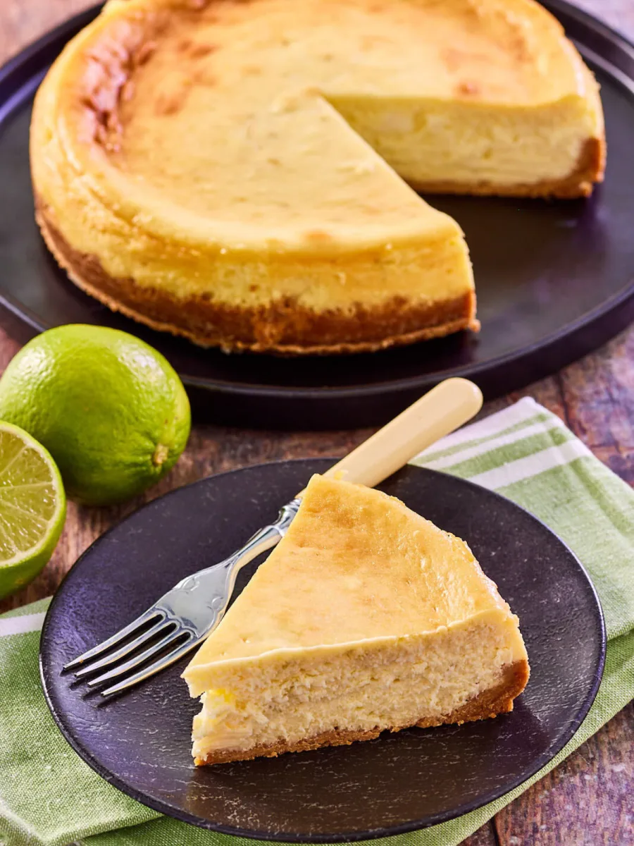 Angled view of a Key Lime Pie Cheesecake slice sitting on a plate with the cut cheesecake in the back - Hostess At Heart