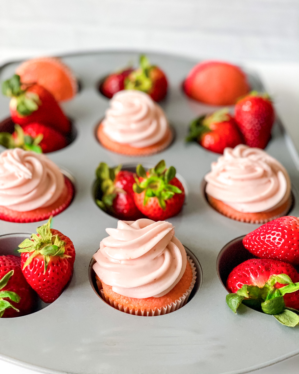 A muffin pan filled with frosted pink cupcakes alternating with fresh strawberries - Hostess At Heart