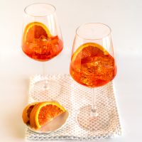 Two orange-hued Aperol Spritz Cocktails garnished with slices of blood orange. Hostess At Heart