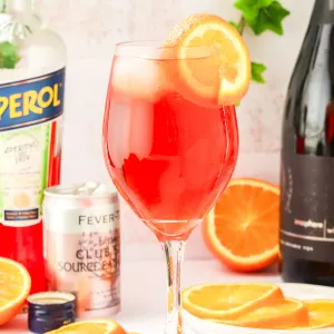 Tableview of a Aperol Spritz garnished with an orange and surrounded by the ingredients. Hostess At Heart