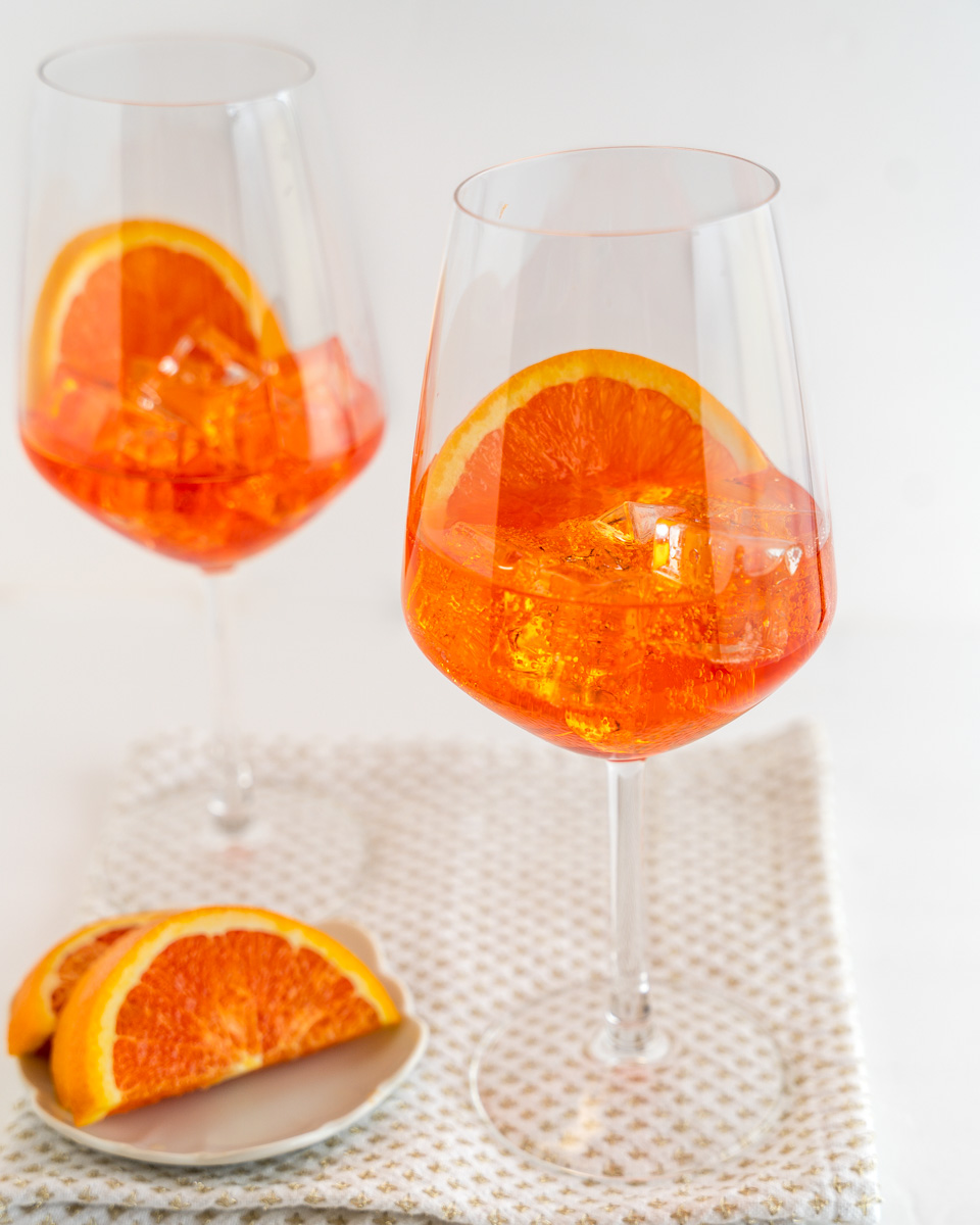 Front view of an orange-hued cocktail in a wine glass next to a plate of sliced blood oranges - Hostess At Heart
