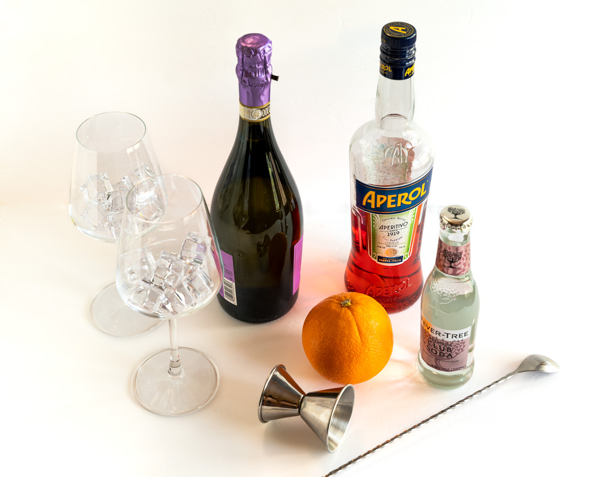 Ingredients used aperol spritzers including prosecco, aperol, and club soda next to two glasses of ice.