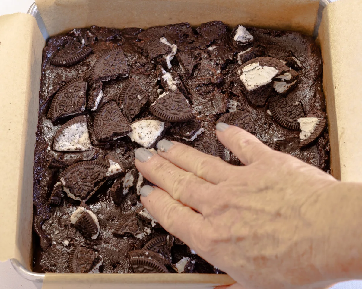 A hand pressing chopped Oreos into chocolate brownie batter - Hostess At Heart