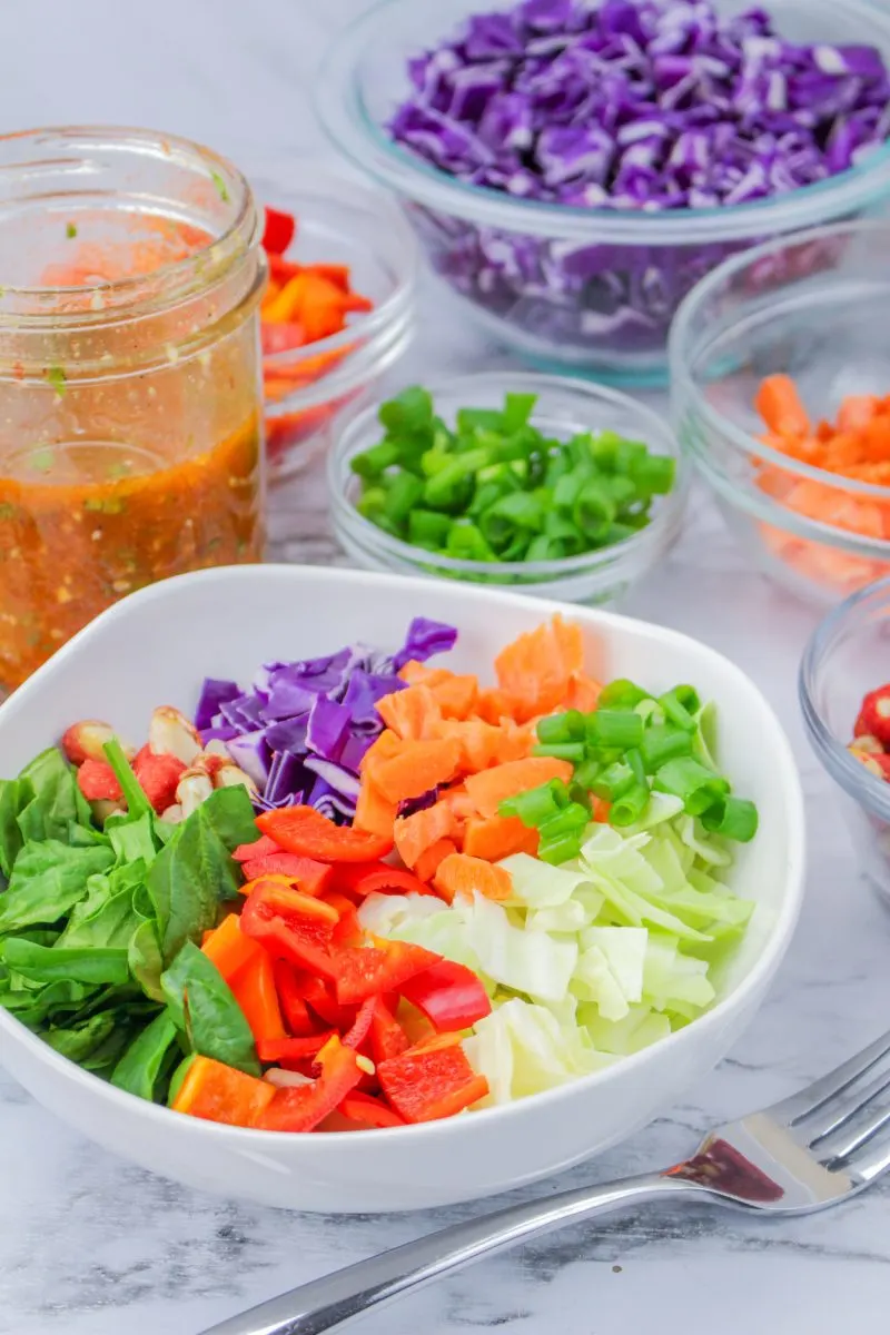 Sideview of a bowl filled with lettuce and chopped vegetables with a jar of dressing and more ingredients in the background - Hostess At Heart