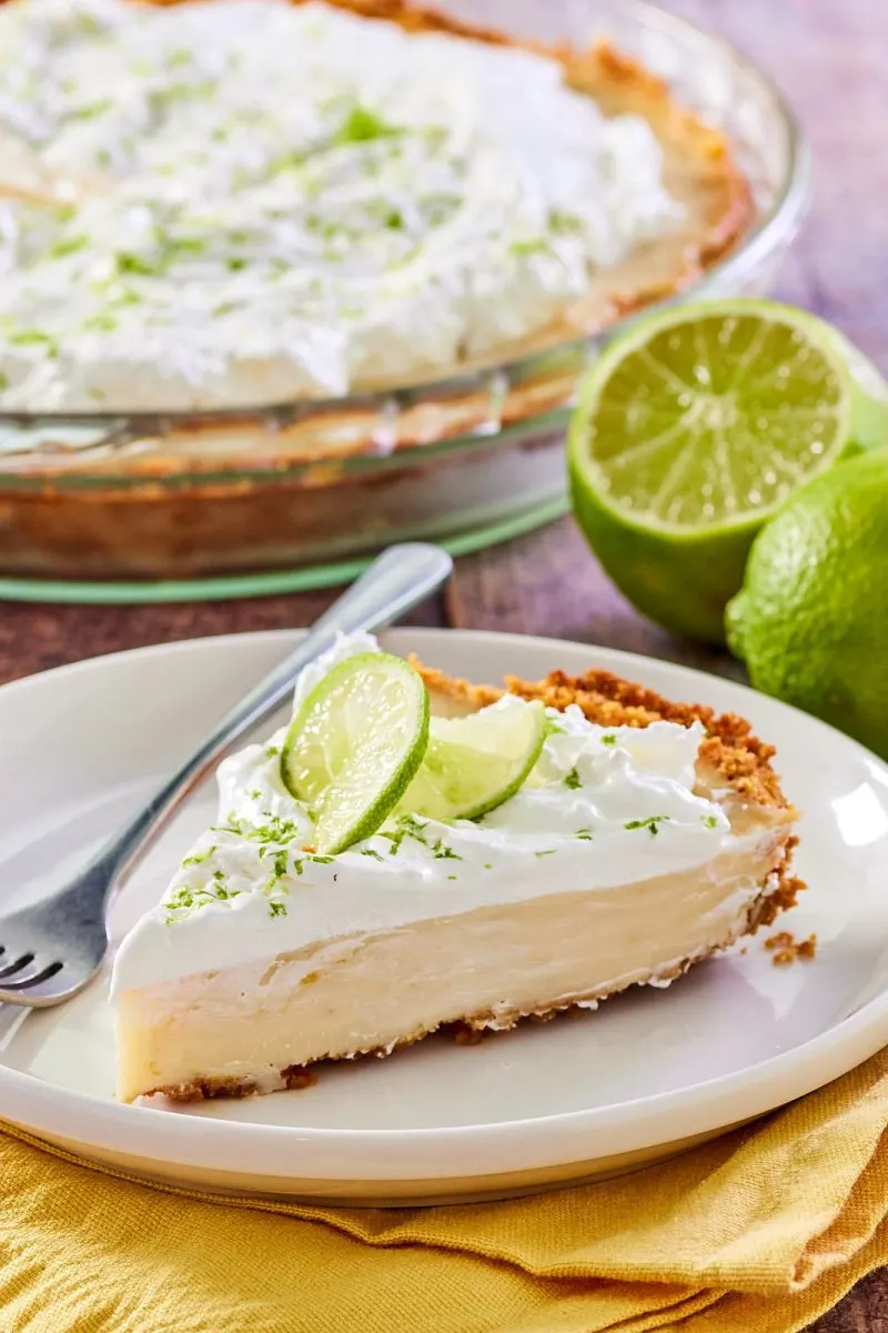 Sideview of a creamy slice of key lime pie in a graham cracker crust garnished with zest and slices of fresh lime.