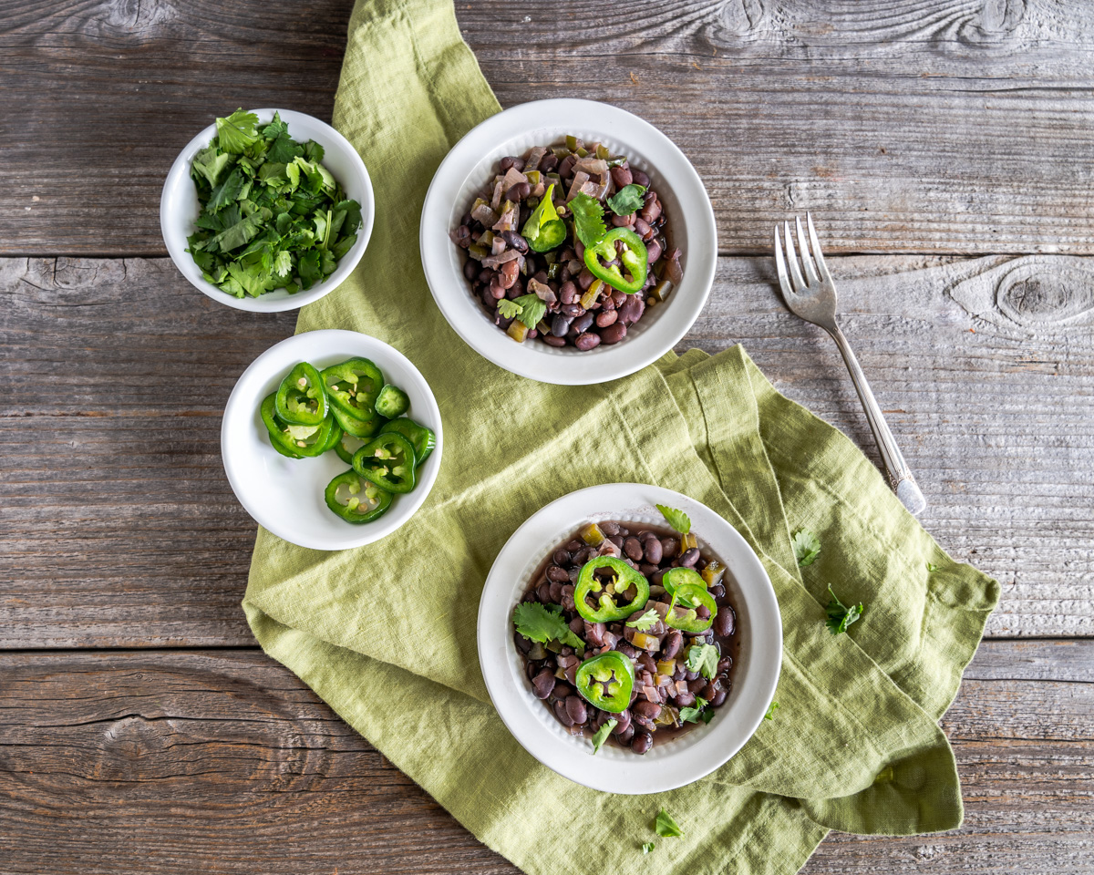 Top Down view of two bowls containing Frijoles Negros next to bowls of cilantro and sliced jalapenos for garnishing - Hostess At Heart