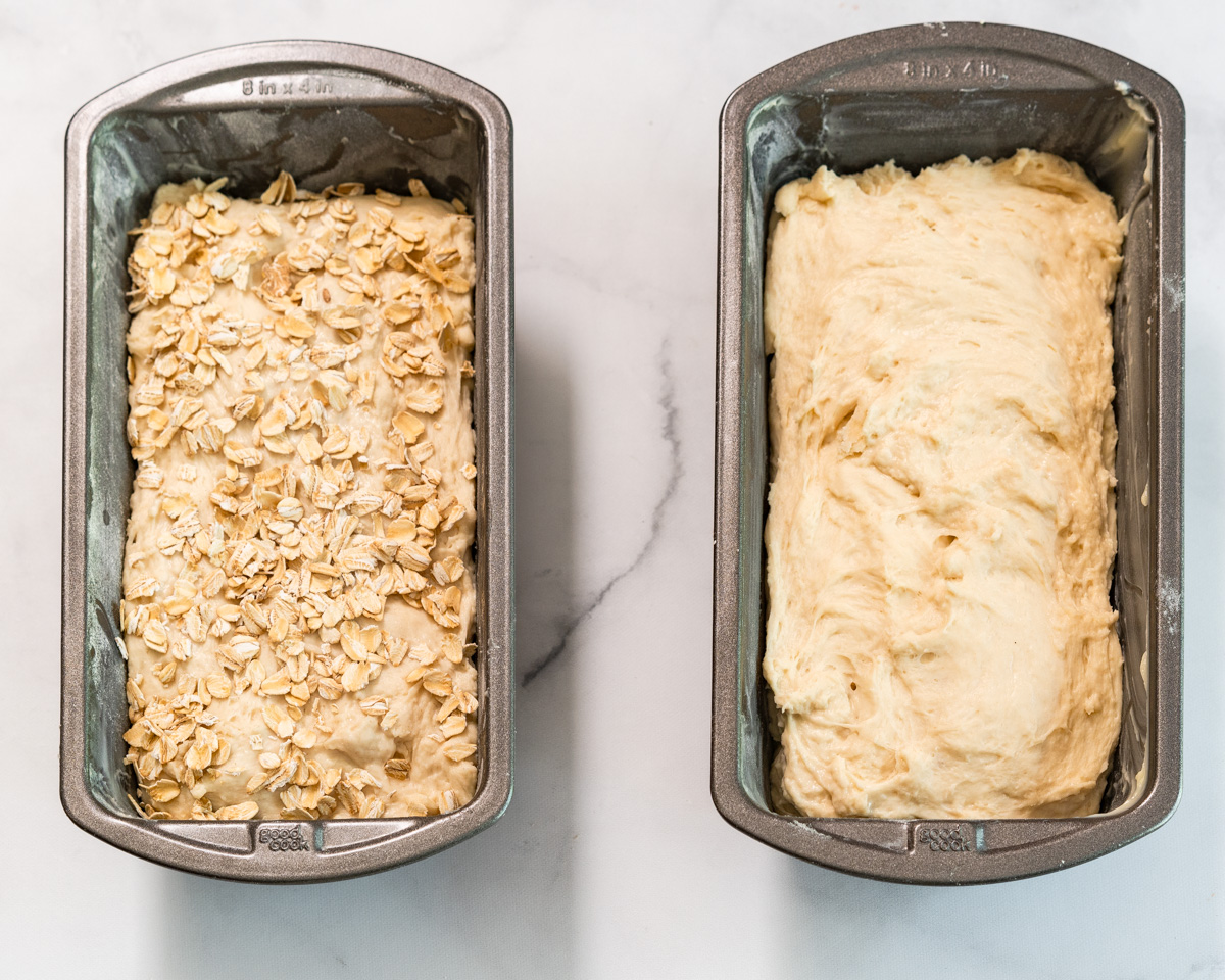 Top down view of two bread pans filled with bread dough before rising. One loaf is topped with oats.