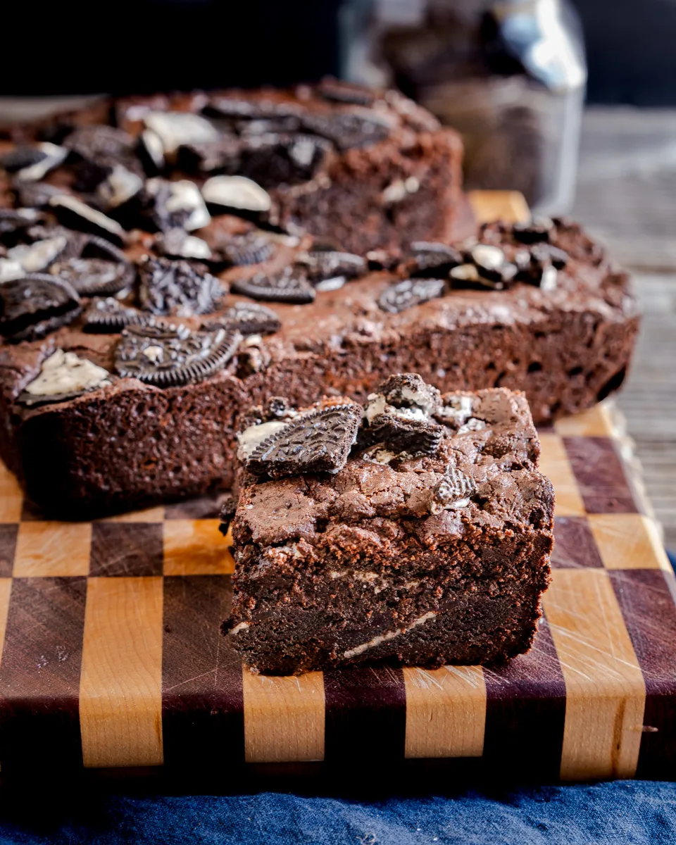 A cutting board with a Oreo Brownie sitting in front of a pan of brownies. Hostess At Heart