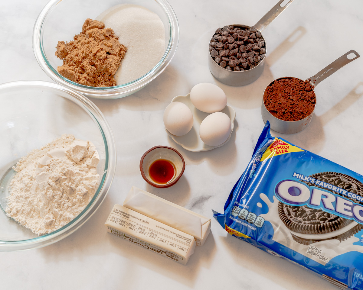 Ingredients used to make Oreo Brownies Recipe including a bag of Oreos, butter, vanilla, flour, cocoa powder, brown sugar, white sugar, eggs, and chocolate chips. Hostess At Heart