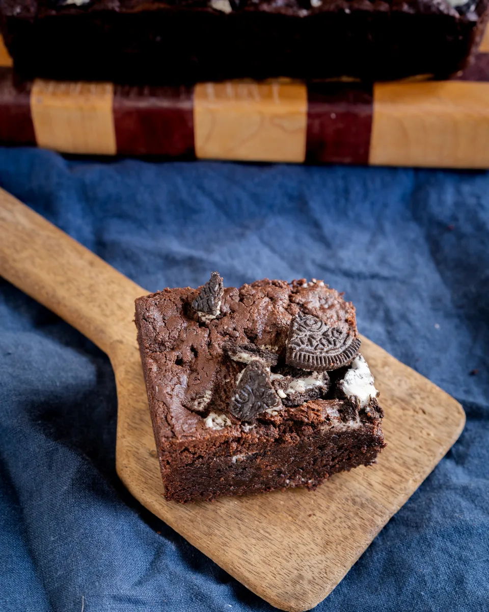 A piece of chocolate brownie stuffed with Oreos sitting on a wooden paddle. - Hostess At Heart