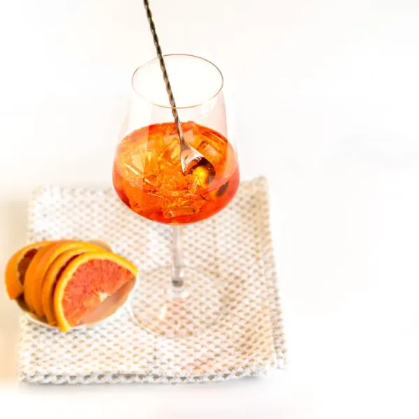 Front view of a glass of apperol spritz sitting next to a dish of blood oranges for garnish - Hostess At Heart