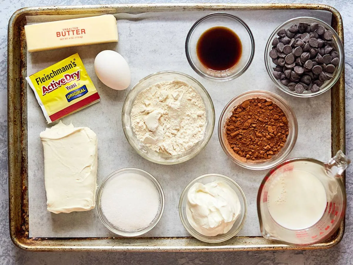 A baking sheet containing babka ingredients including flour, sugar, farmers cheese, cocoa, egg, butter, yeast, chocolate chips, vanilla, milk, and sour cream.