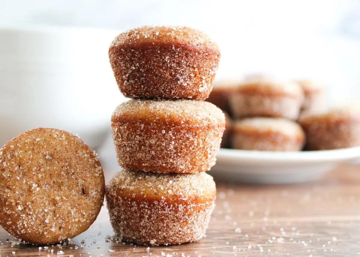 A sideview of baked mini Apple Cider Donut Muffins stacked on top of each other with one muffin laying on it's side and several more muffins on a platter in the back ground - Hostess At Heart