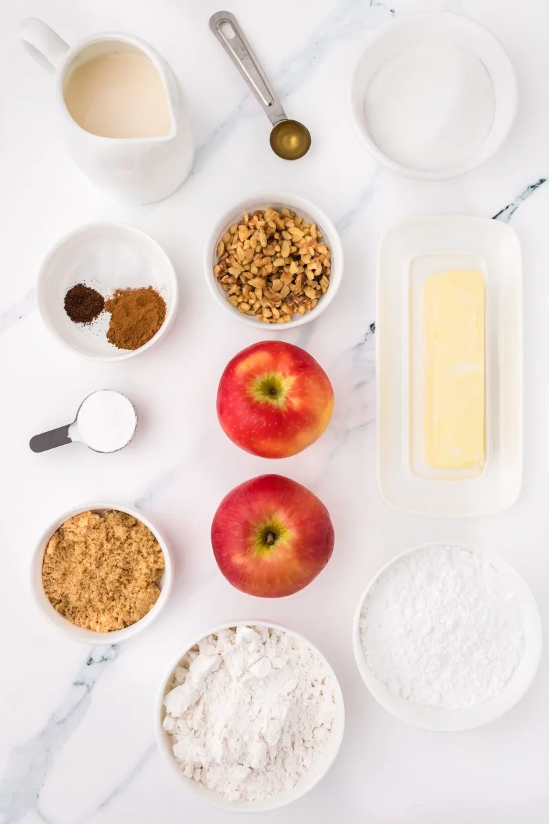 Top down view of individual dishes holding the ingredients need to make Apple Muffins with Crumble Topping including apples, chopped nuts, apple cider vinegar, flour, white sugar, butter, cinnamon, cloves, powdered sugar, baking powder, and milk. Hostess At Heart.