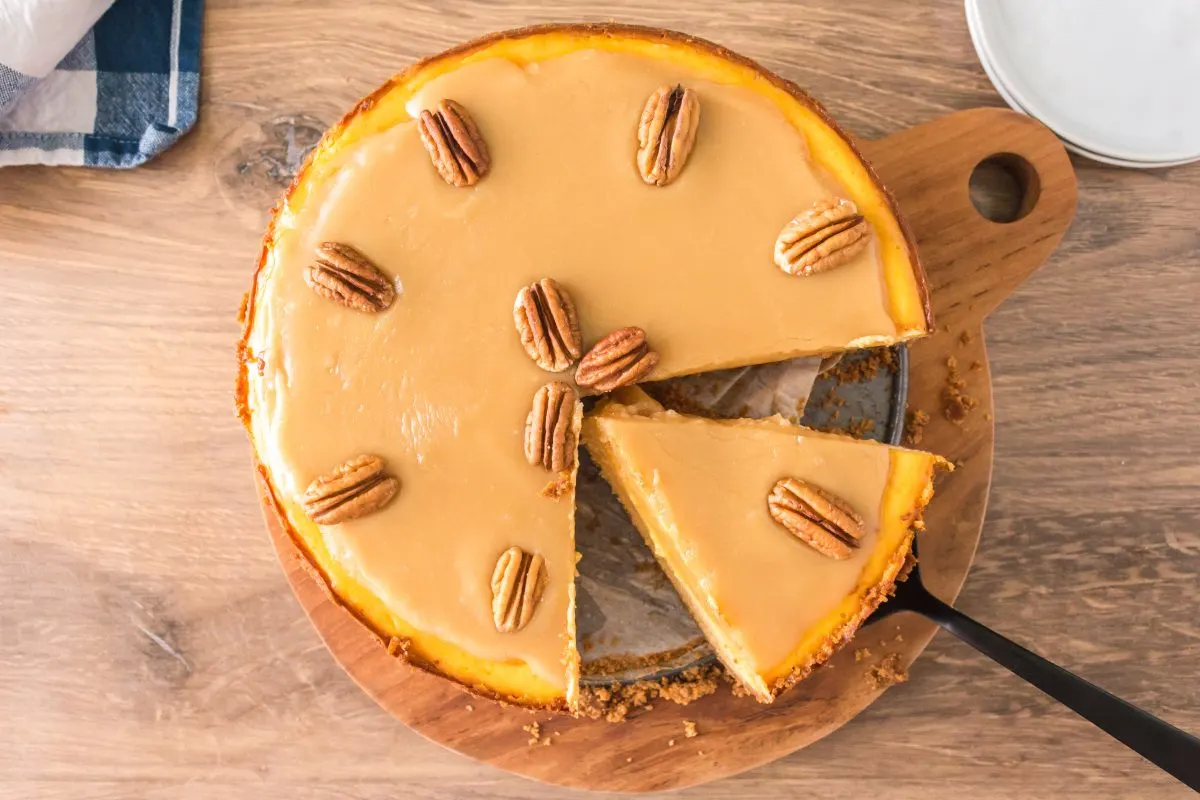 Top down view of a praline cheesecake topped with pecans with one slice being lifted out with a pie server - Hostess At Heart