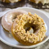 Two homemade doughnuts on a plate. Both dipped in vanilla icing and one topped with crushed nuts - Hostess At Heart