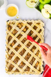Top down view of egg wash being applied to a sheet pan apple caramel pie with lattice crust - Hostess At Heart